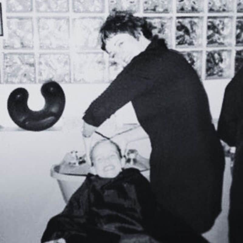 A person is washing the hair of another person, who is seated in a salon chair, both smiling. The scene takes place in a salon with a glass block wall in the background.