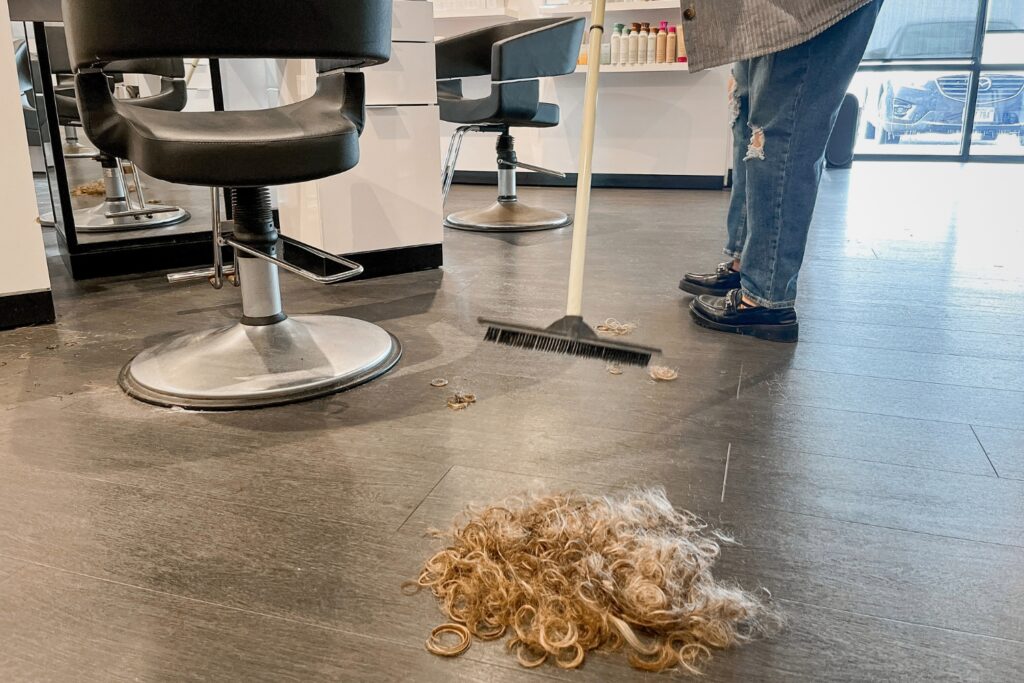 A person sweeps clumps of hair from the floor in a salon next to an empty salon chair.