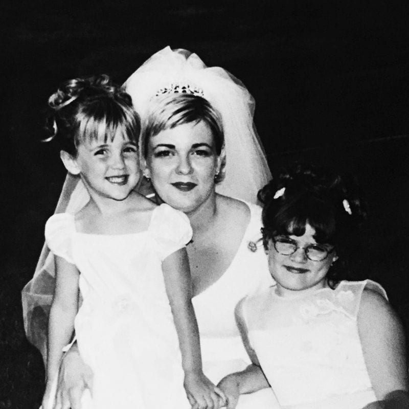 A bride wearing a veil and tiara poses for a photo with two young girls in white dresses, one smiling broadly and the other wearing glasses.