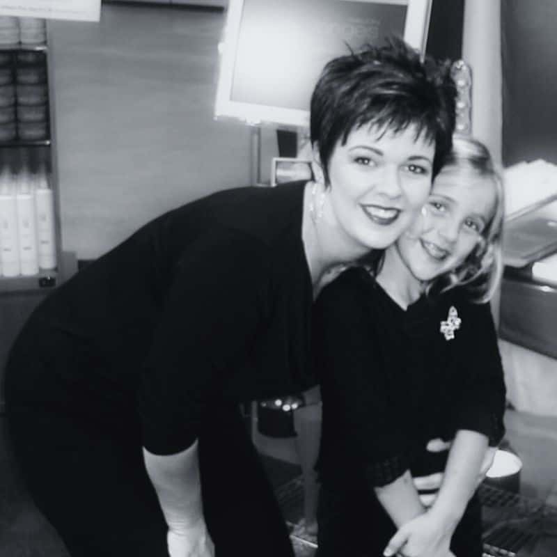 A woman with short hair smiles while leaning down to hug a young girl with long hair. Both are wearing dark clothing and posing in an indoor setting.