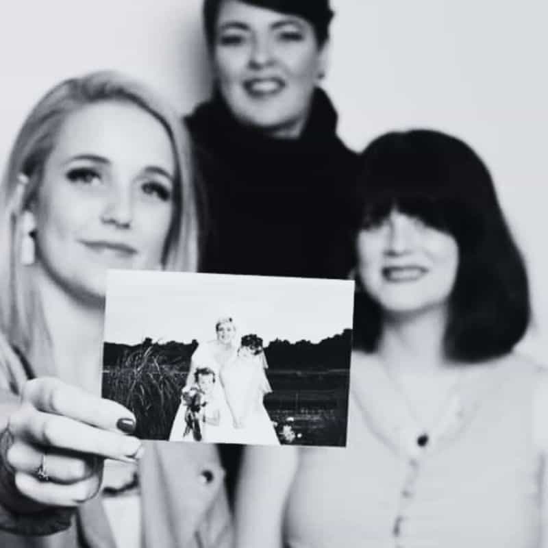 Three women posing for a black and white photo, with one holding a picture of a bride and groom taken outdoors.