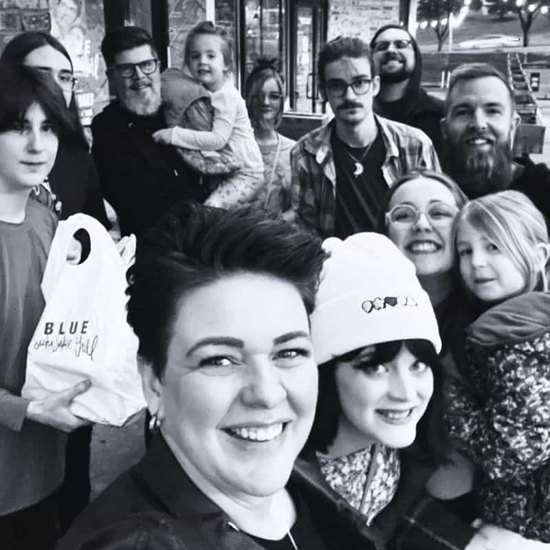 A group of people, including adults and children, pose closely together for a selfie outside a building. Some are smiling, and one person is holding a bag with the text "BLUE". The image is in black and white.