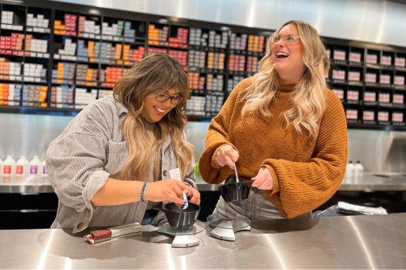 Two women in a hair salon mix hair color in bowls, smiling and laughing. Behind them, shelves filled with various hair dye products add a splash of vibrant colors to the friendly atmosphere.