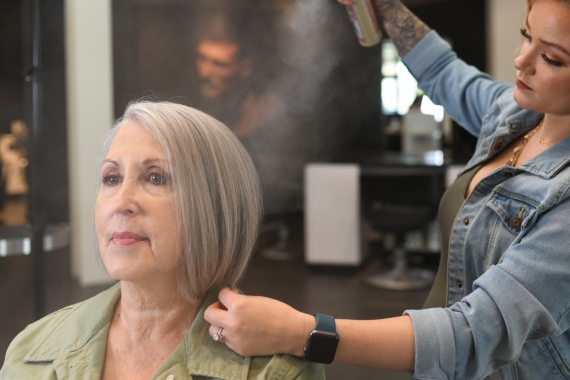 A hairstylist in a denim jacket sprays a middle aged woman’s short gray hair at the hair salon. The woman, wearing a green shirt, sits comfortably in the salon chair, her new look taking shape.