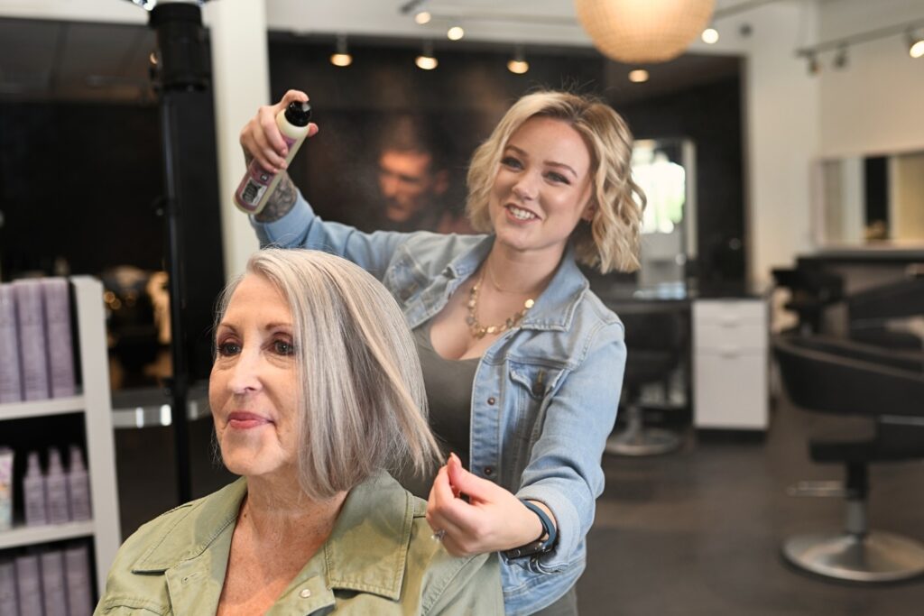 A hairstylist sprays a product on the hair of an older woman with gray hair in a salon. The stylist smiles while working, and the stylish salon interior is visible in the background.
