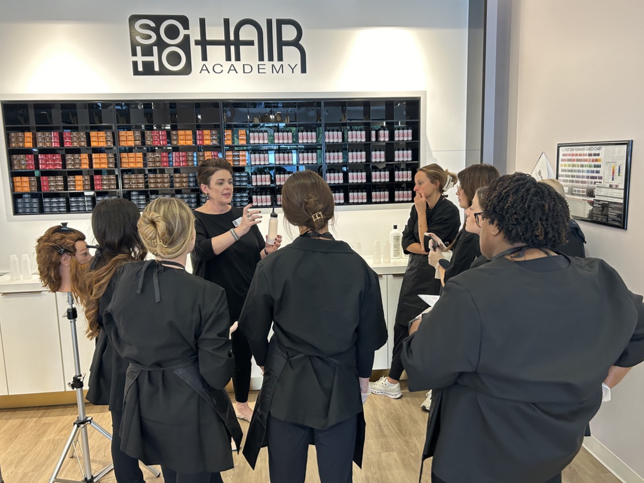 A group of students in black uniforms gather around an instructor in a hair academy, with shelves of hair products in the background.