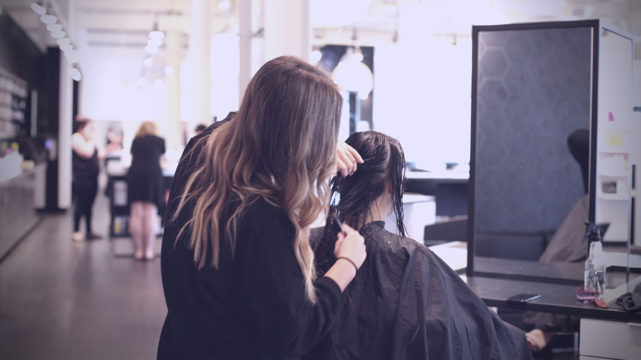 A hairstylist is cutting a client's hair in a salon, with more clients and stylists visible in the background.