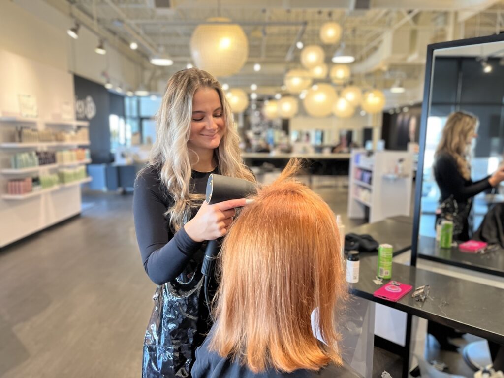 Hair stylist blow drying a client's hair in a modern salon with shelves of products and round ceiling lights in the background.