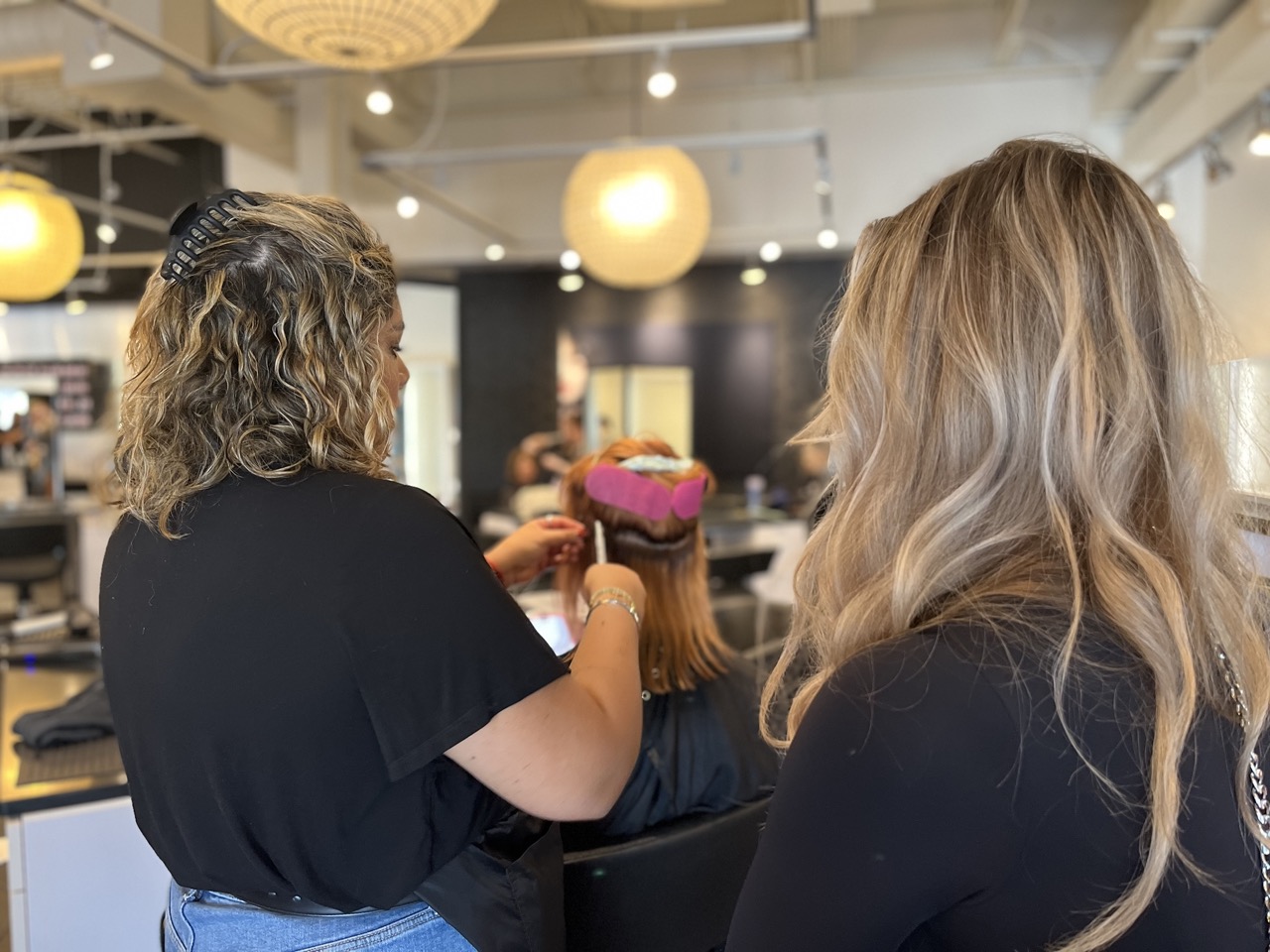 A hairstylist applies color to a client's hair while another person observes in a salon.