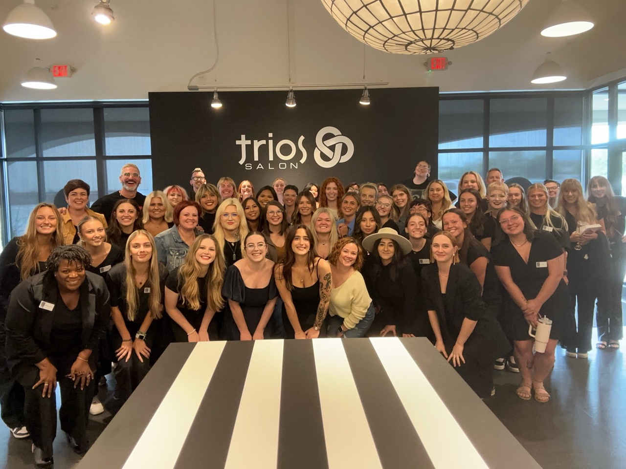 A large group of people posing in front of a sign that reads "trios salon" indoors.