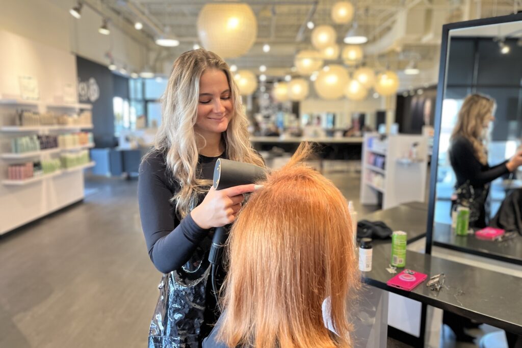 A skilled stylist blow dries a client's red hair in a modern salon. The bright interior, featuring large round lights and shelves stocked with top notch hair products, creates the perfect ambiance for styling.