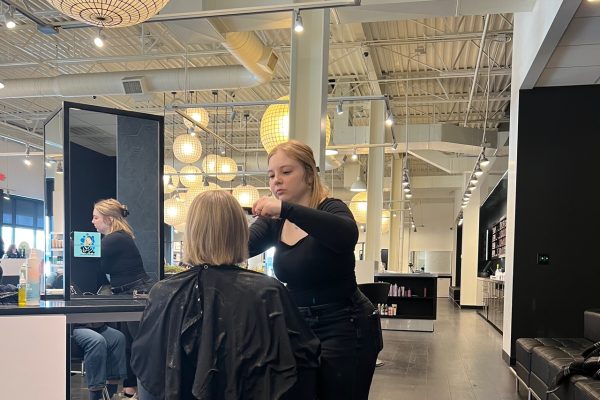 A stylist cuts hair at a salon, with a client seated in a chair. The salon has large illuminated spherical lamps and a modern design.