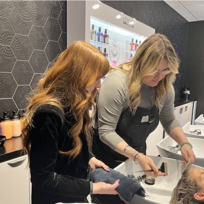 Two hairstylists work in a hair salon, with one washing a client's hair at a sink and the other holding a towel. Shelves brimming with top notch salon products are visible in the background.